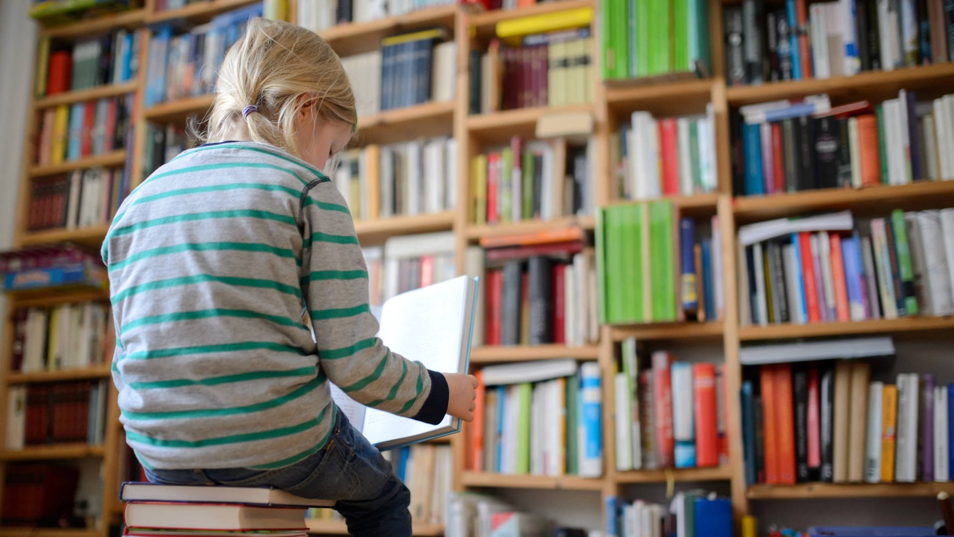 Ein Kind sitzt auf einem Buchstapel und liest ein Buch, im Hintergrund ein Bücherregal.