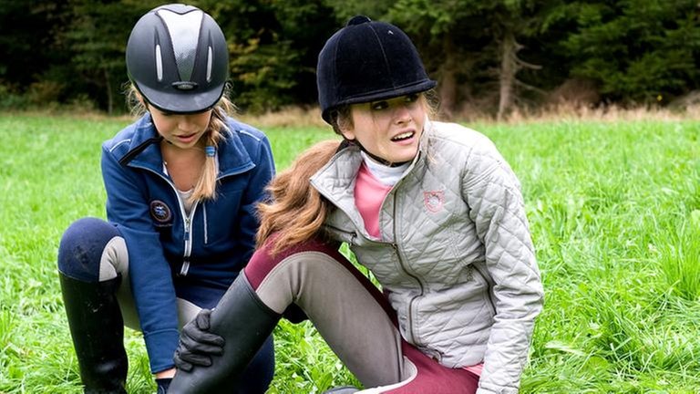 Zwei Mädchen in Reitkleidung sitzen auf einer Wiese, die eine hält sich ihr rechtes Bein (Foto: SWR, SWR -)