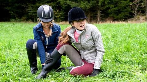Zwei Mädchen in Reitkleidung sitzen auf einer Wiese, die eine hält sich ihr rechtes Bein (Foto: SWR, SWR -)