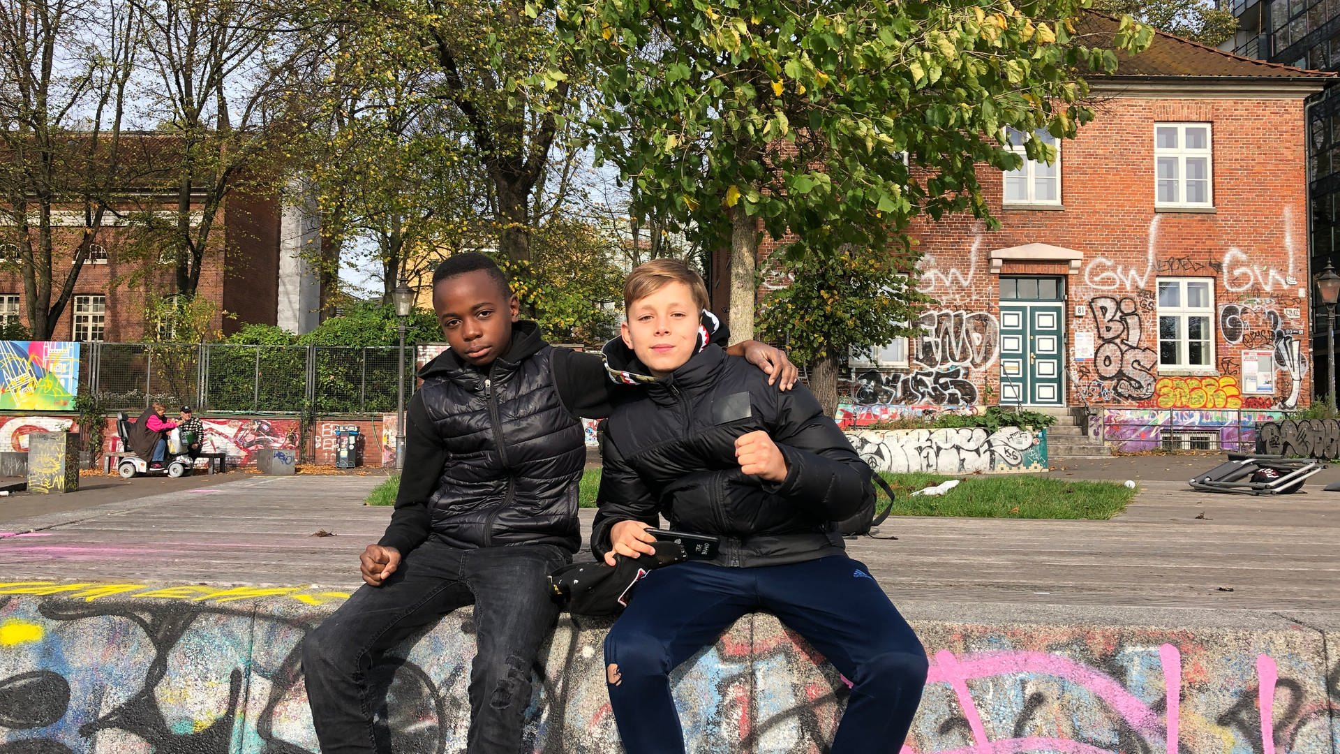 Lionnel (l.) und Otto (r.) sitzen in einem Park vor mit Graffiti bemalten Wänden (Foto: SWR)
