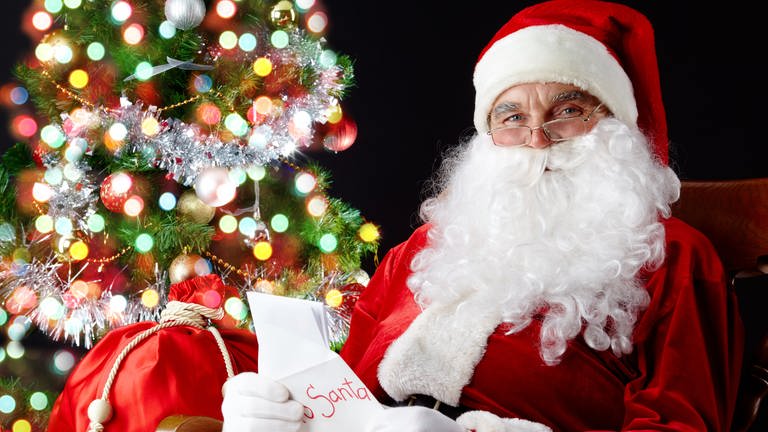 Der Weihnachtsmann vor einem Weihnachtsbaum mit einem Sack Geschenke und einer Weihnachtskarte (Foto: Colourbox)