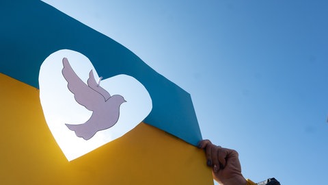 Eine Taube aus Papier schwebt während einer Demonstration gegen den russischen Angriff auf die Ukraine in einer Ukraineflagge aus Papier. (Foto: picture-alliance / Reportdienste, Sebastian Gollnow)