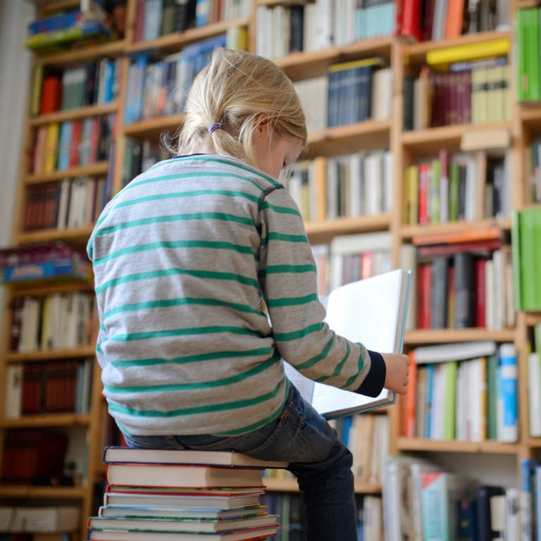 Ein Kind sitzt auf einem Buchstapel und liest ein Buch, im Hintergrund ein Bücherregal.