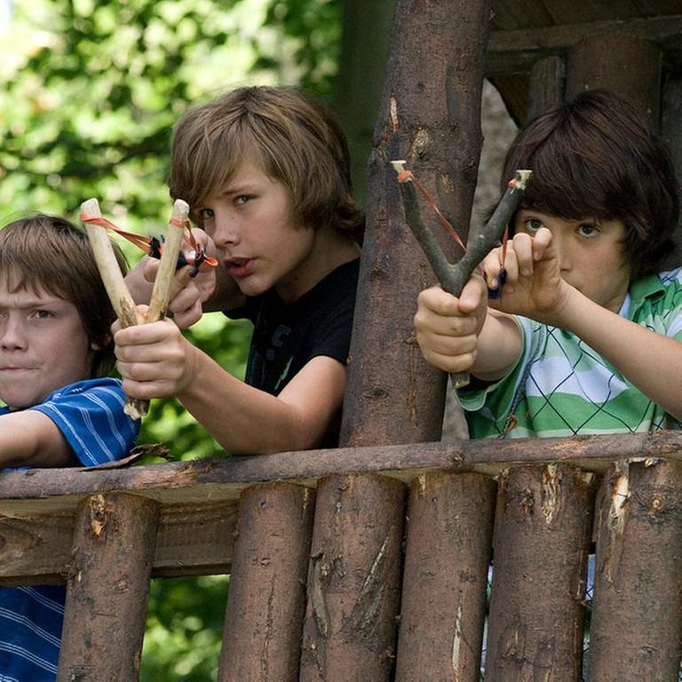 Drei Jungs stehen in ihrem Baumhaus und haben Steinschleudern gespannt zum Abschuss